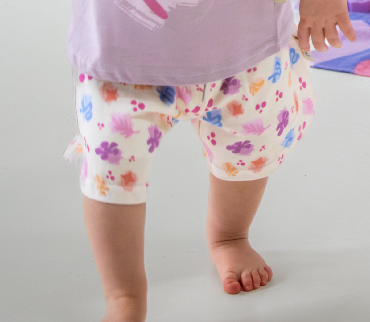 Child wearing a purple shirt with pink leaf design and floral shorts, standing on a light gray background.