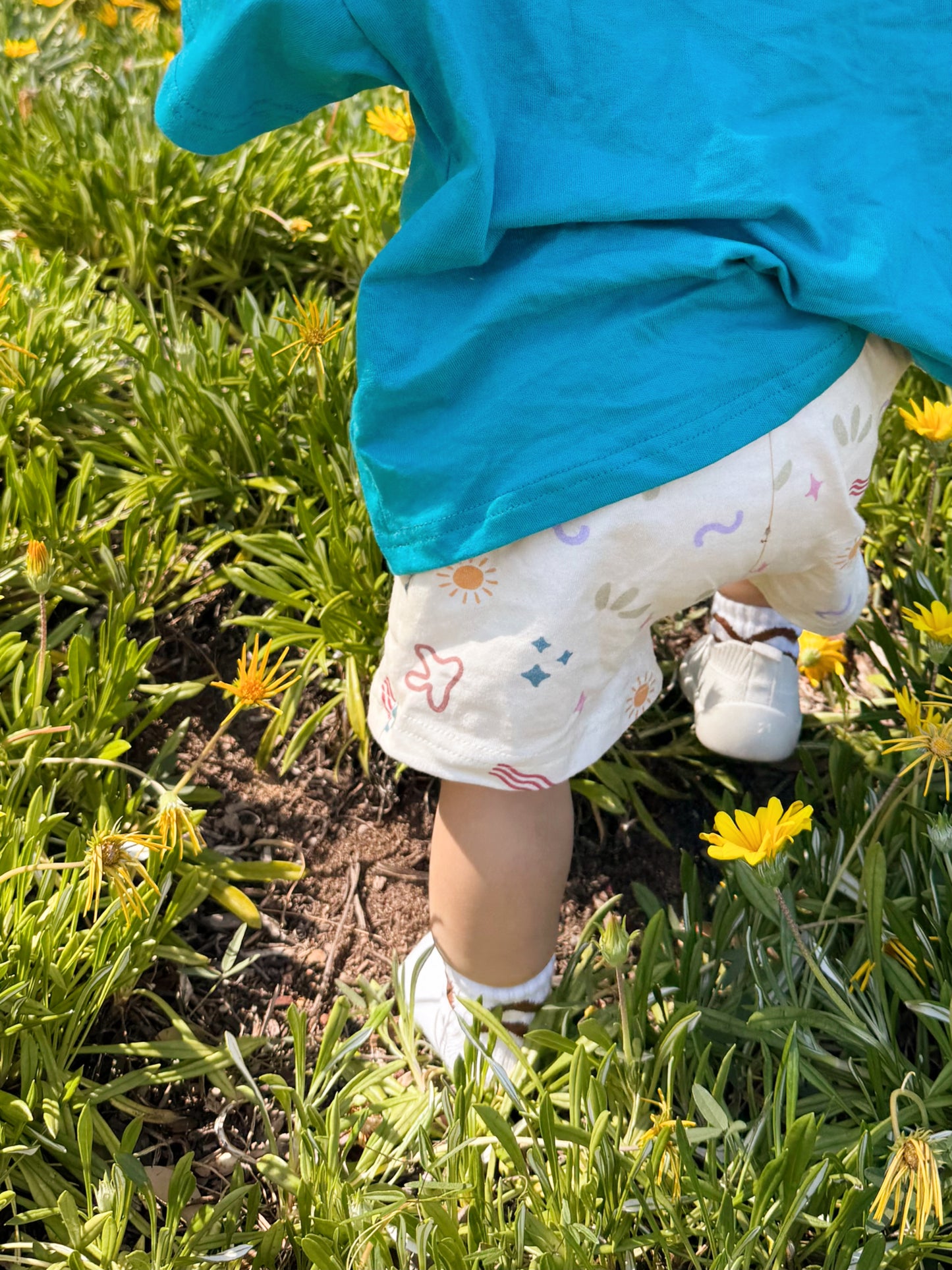 Child in a blue shirt and patterned shorts standing in grass with yellow flowers
