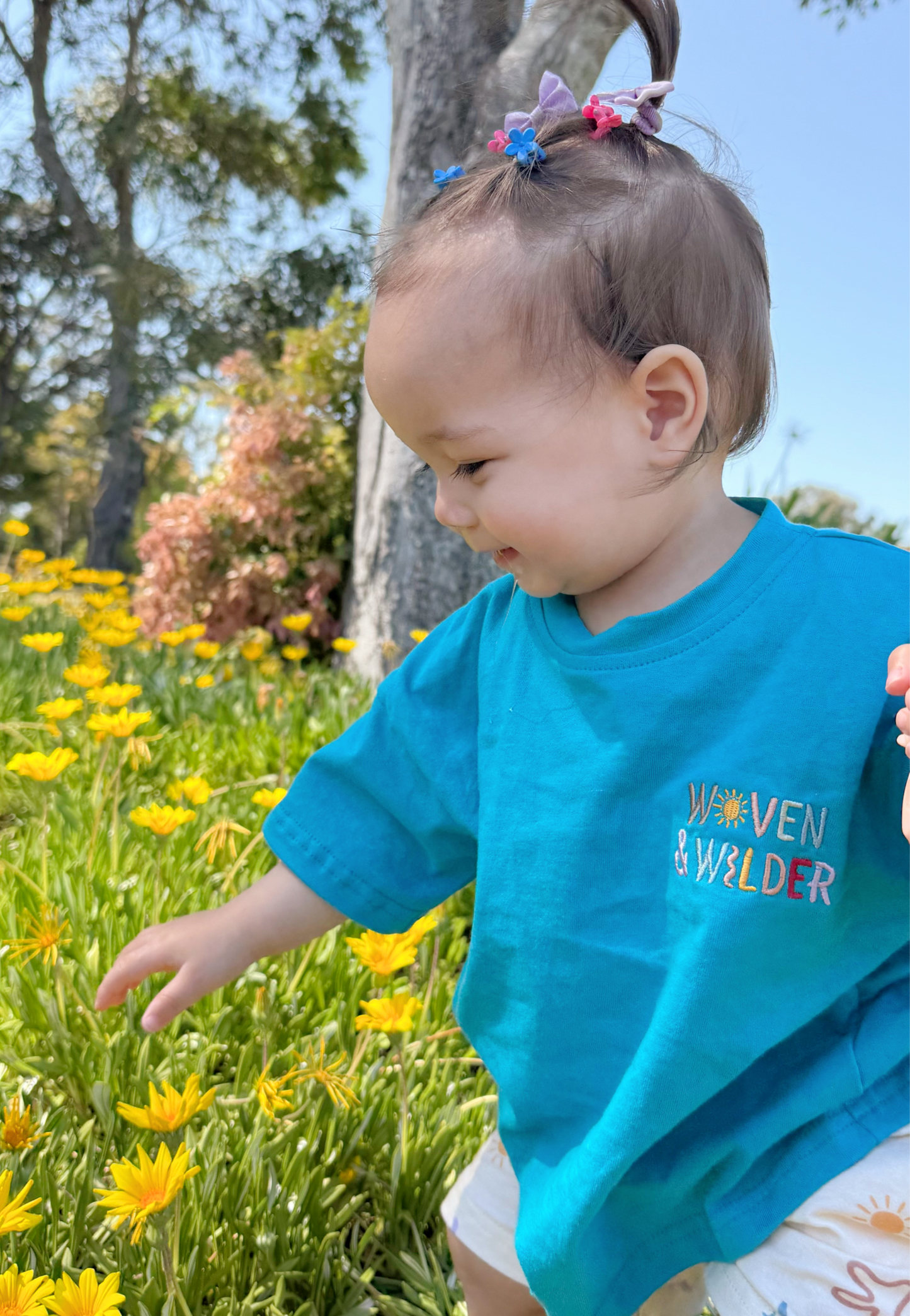 Child in a blue shirt with 'Woven & Wilder' text standing in a field of yellow flowers.