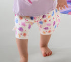 Child wearing a purple shirt with pink leaf design and floral shorts, standing on a light gray background.