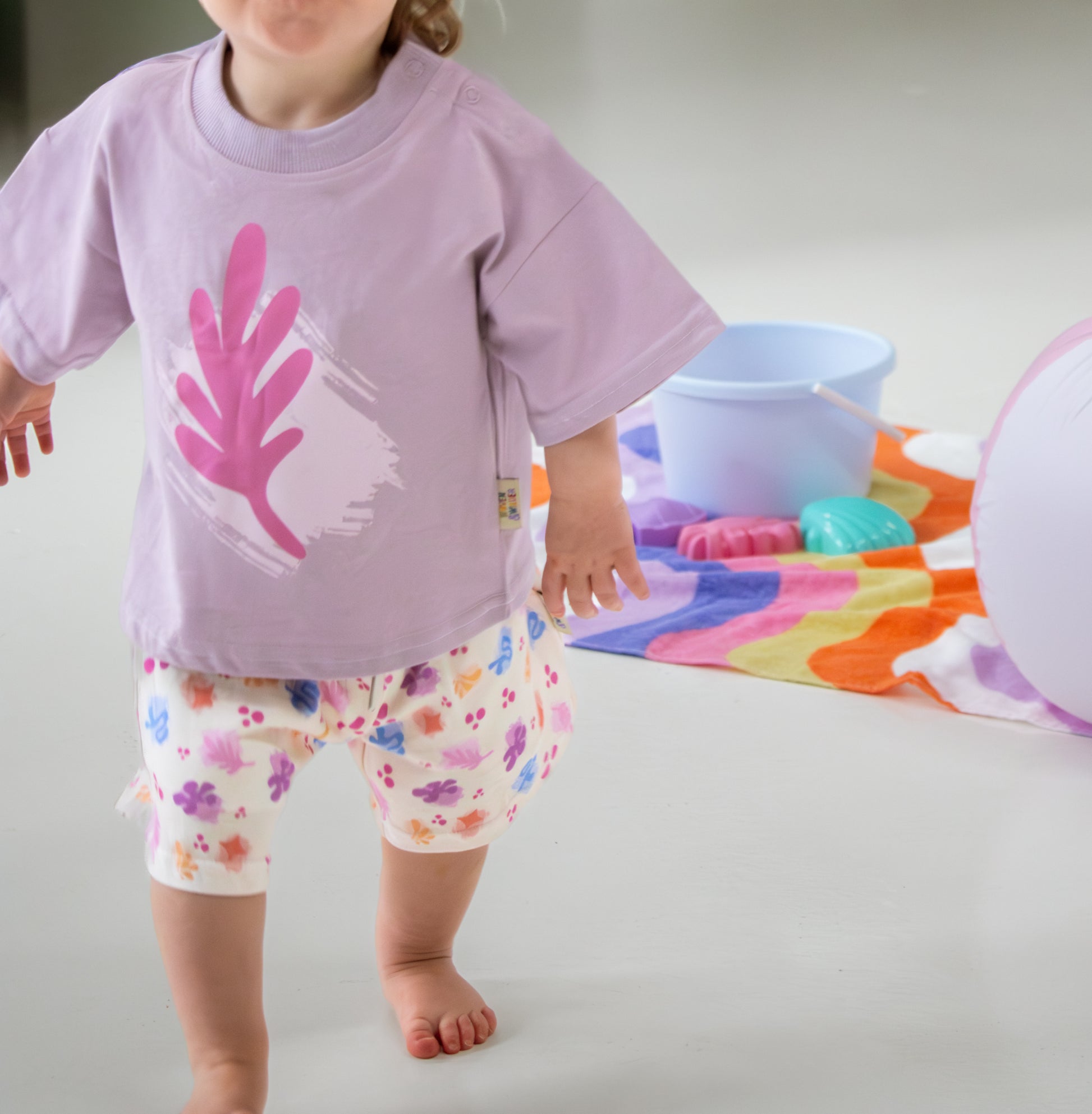 Child wearing a purple shirt with pink leaf design and floral shorts, standing on a light gray background.