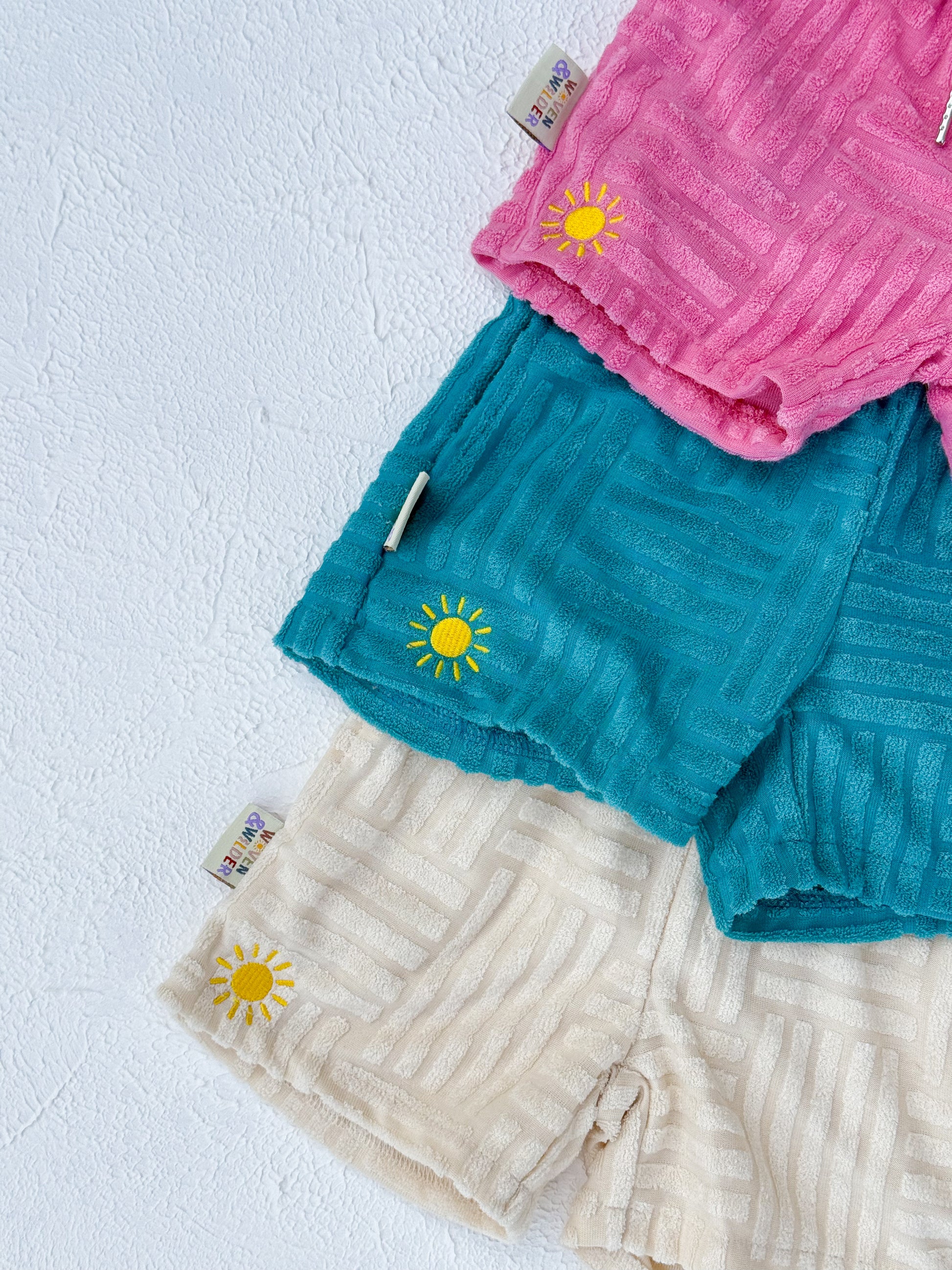 Three baby shorts in pink, blue, and beige with sun designs on a light blue background.