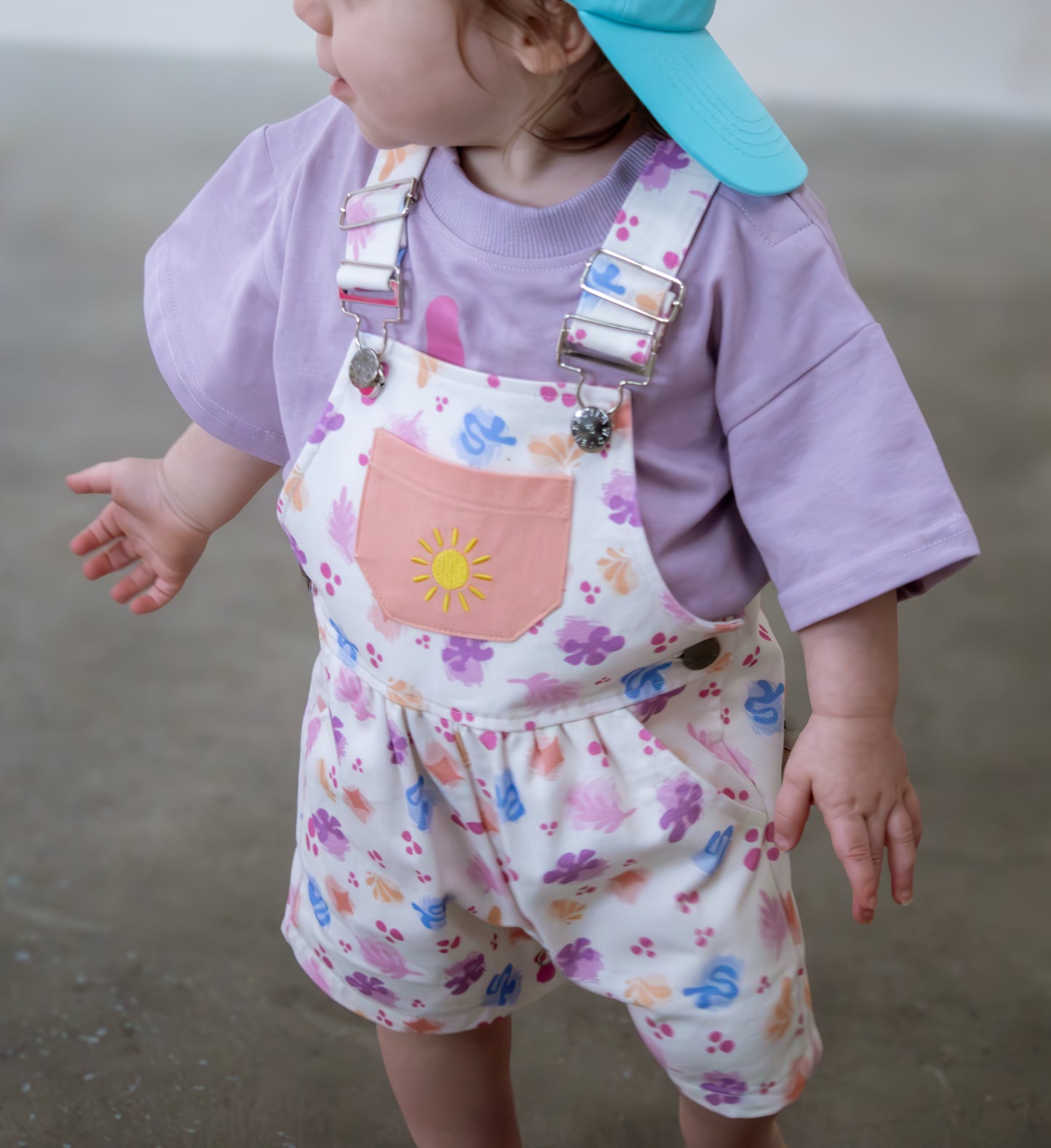 Child wearing a colorful floral romper with a sun design and a purple shirt.