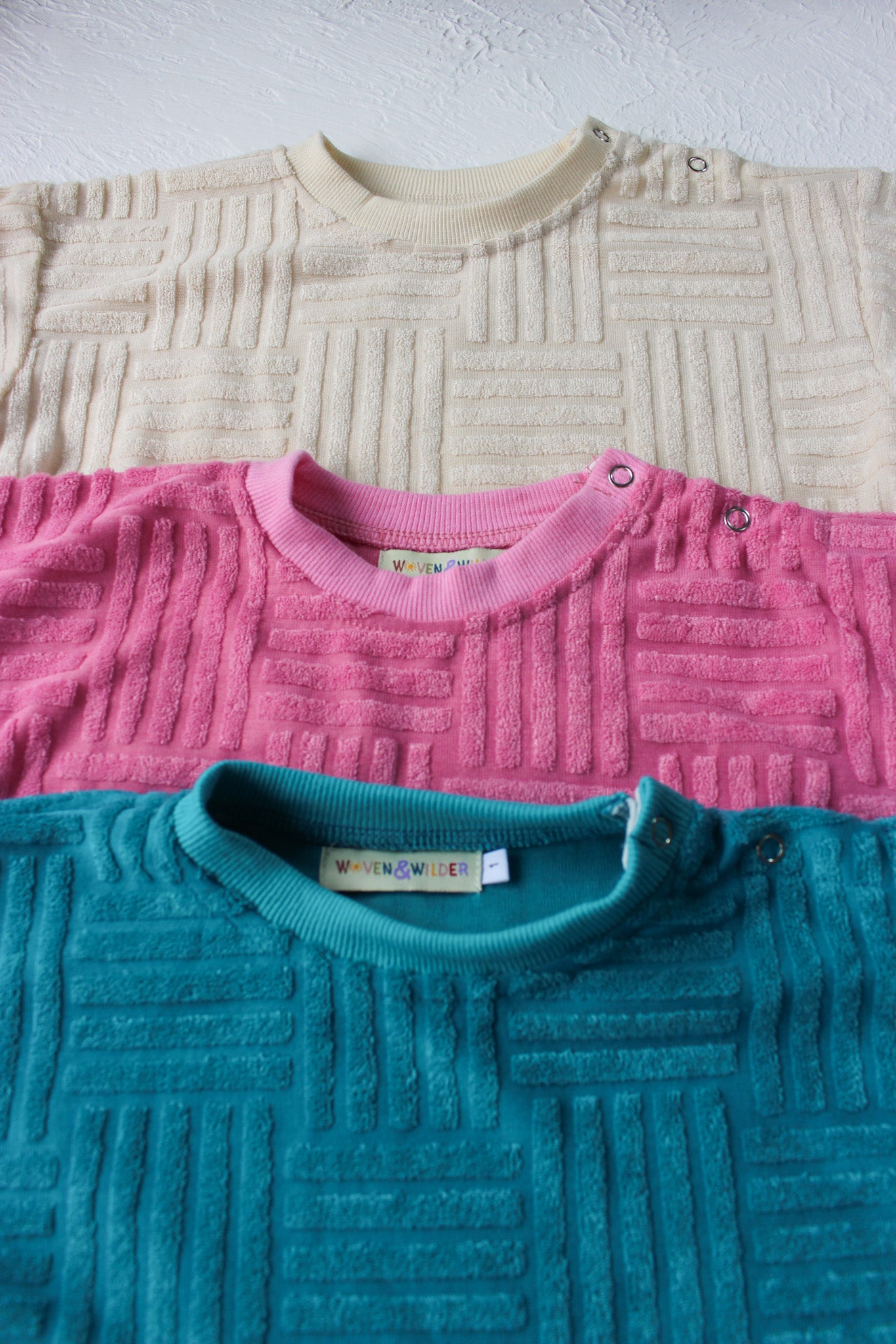 Three textured tshirts in beige, pink, and teal on a light gray background.