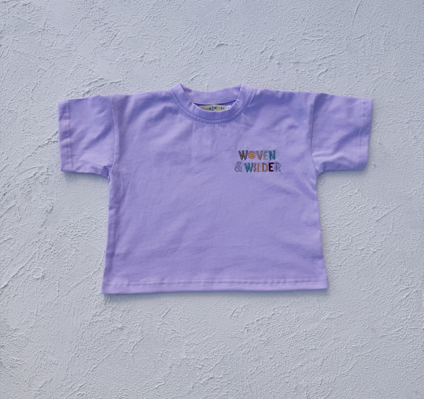 Purple t-shirt with 'Woven & Wilder' text on a light gray background
