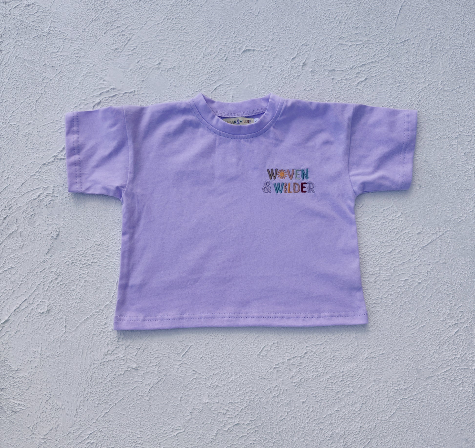 Purple t-shirt with 'Woven & Wilder' text on a light gray background