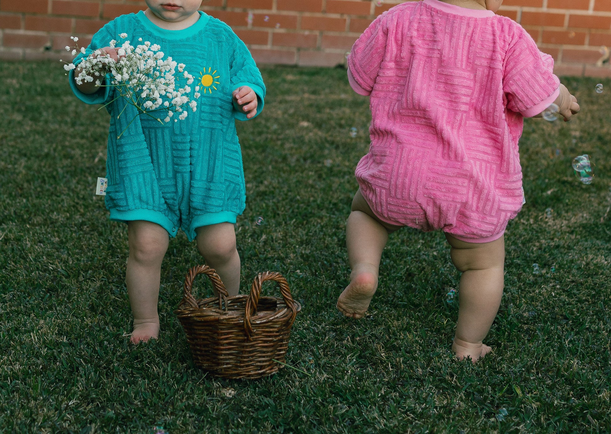 Two children in colorful rompers standing on grass with a basket and flowers.