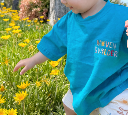 Child in a blue shirt with 'Woven & Wilder' text standing in a field of yellow flowers.