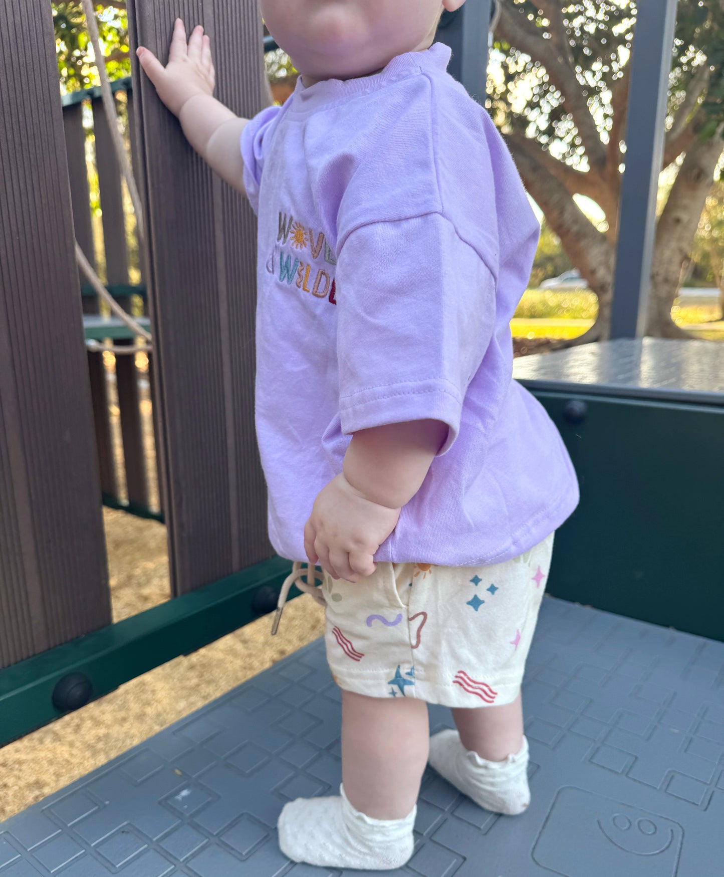 Child in a purple shirt and patterned shorts standing on a playground surface.