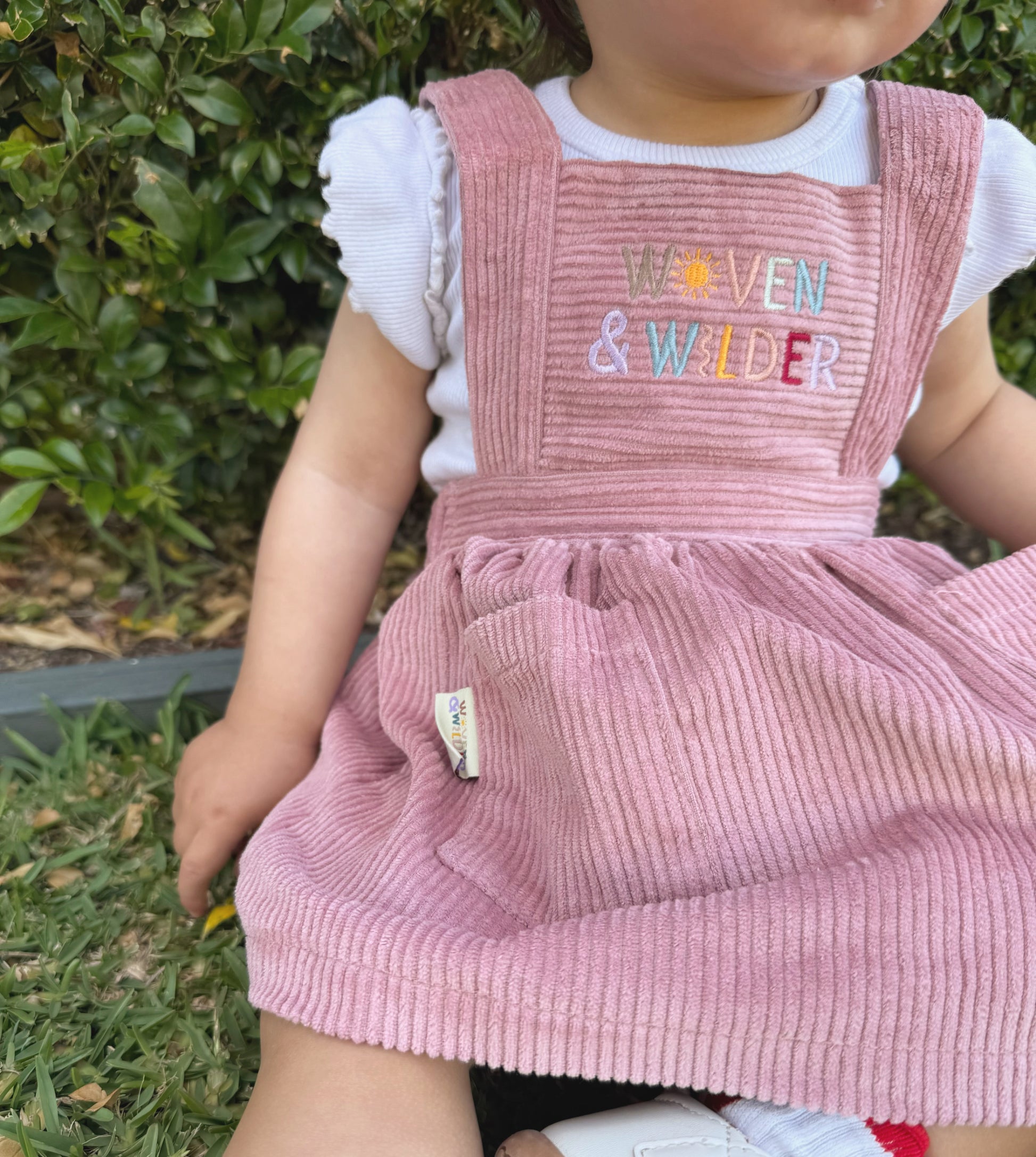 Child wearing a pink corduroy dress with 'Woven & Wilder' embroidery, sitting outdoors.