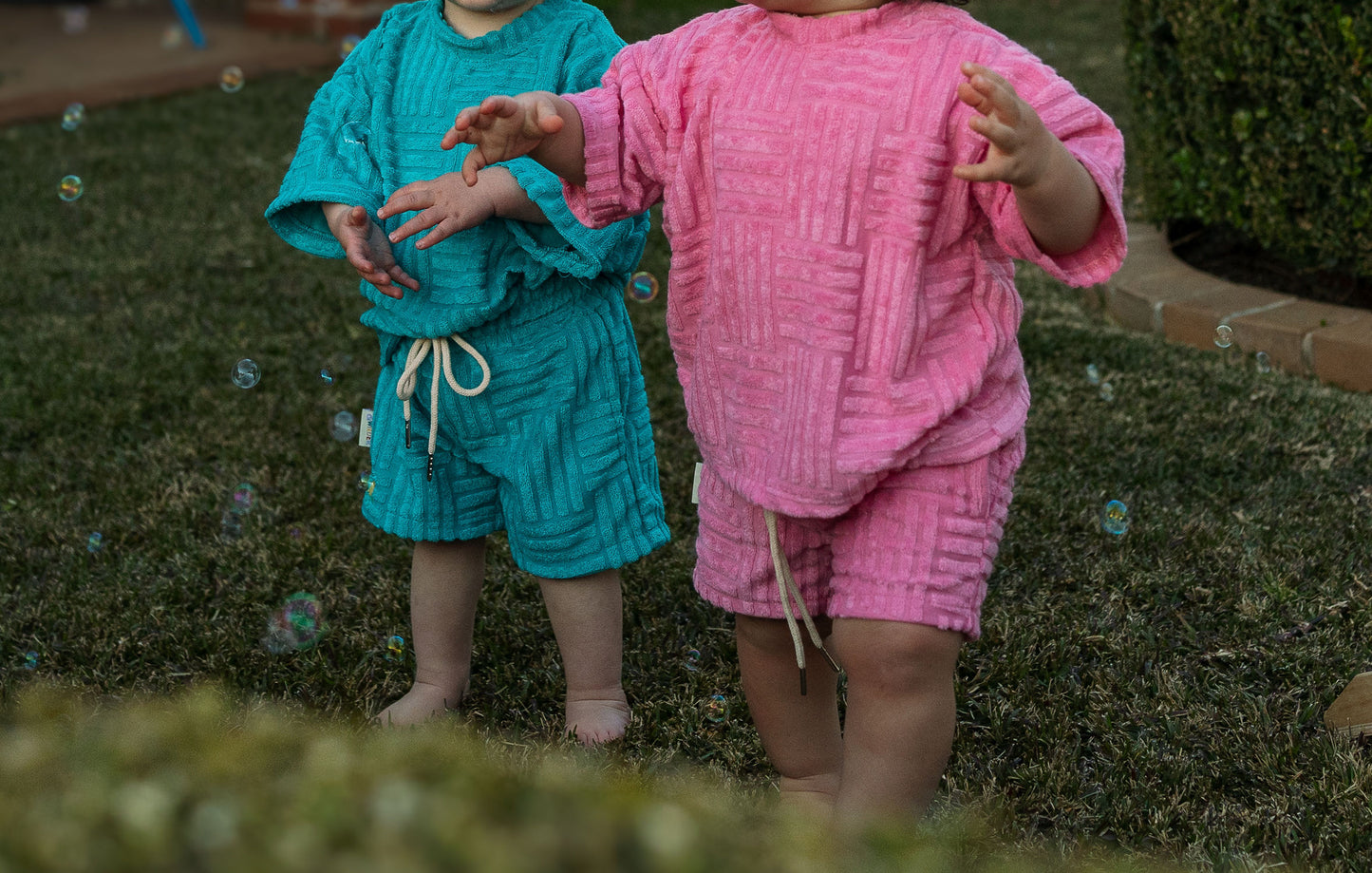 Two children in colorful tshirts standing on grass with bubbles around them.
