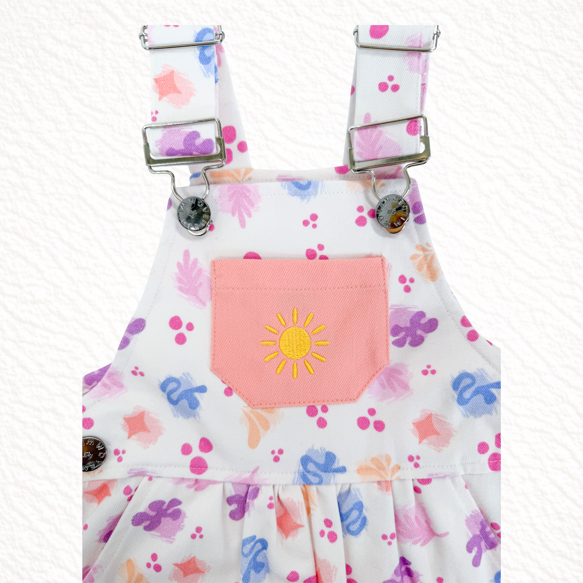 Children's overalls with colorful floral pattern and pink pocket featuring a yellow sun design on a white background.