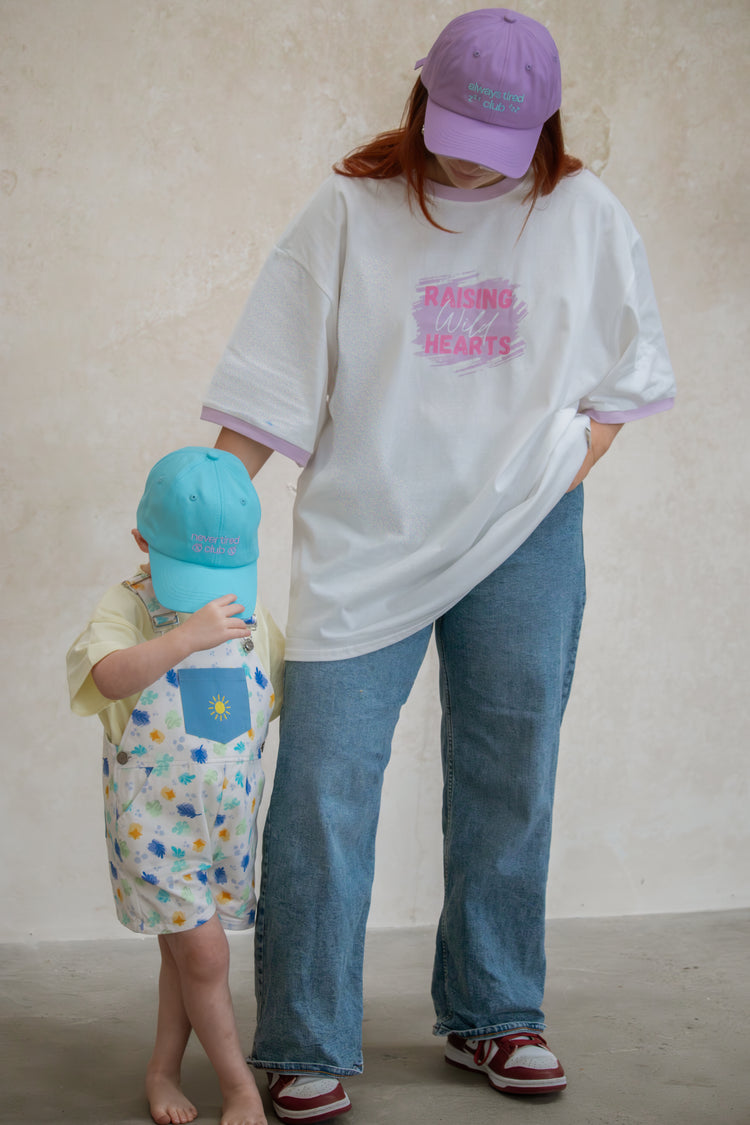 A mother wearing a raising heart tee with her kid wearing an overall with colorful caps against a plain background
