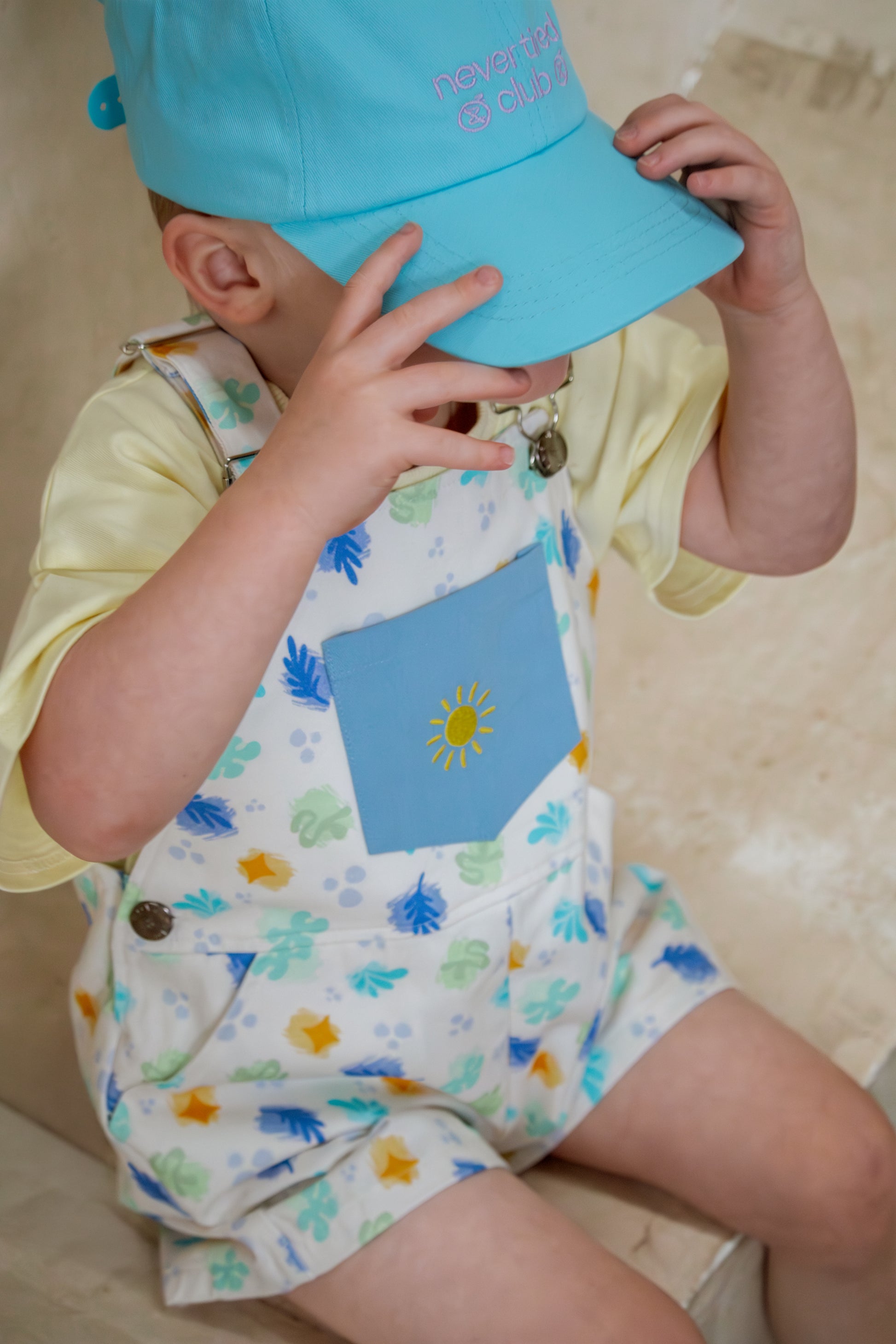 Child wearing a light blue cap and colorful outfit with a sun design.