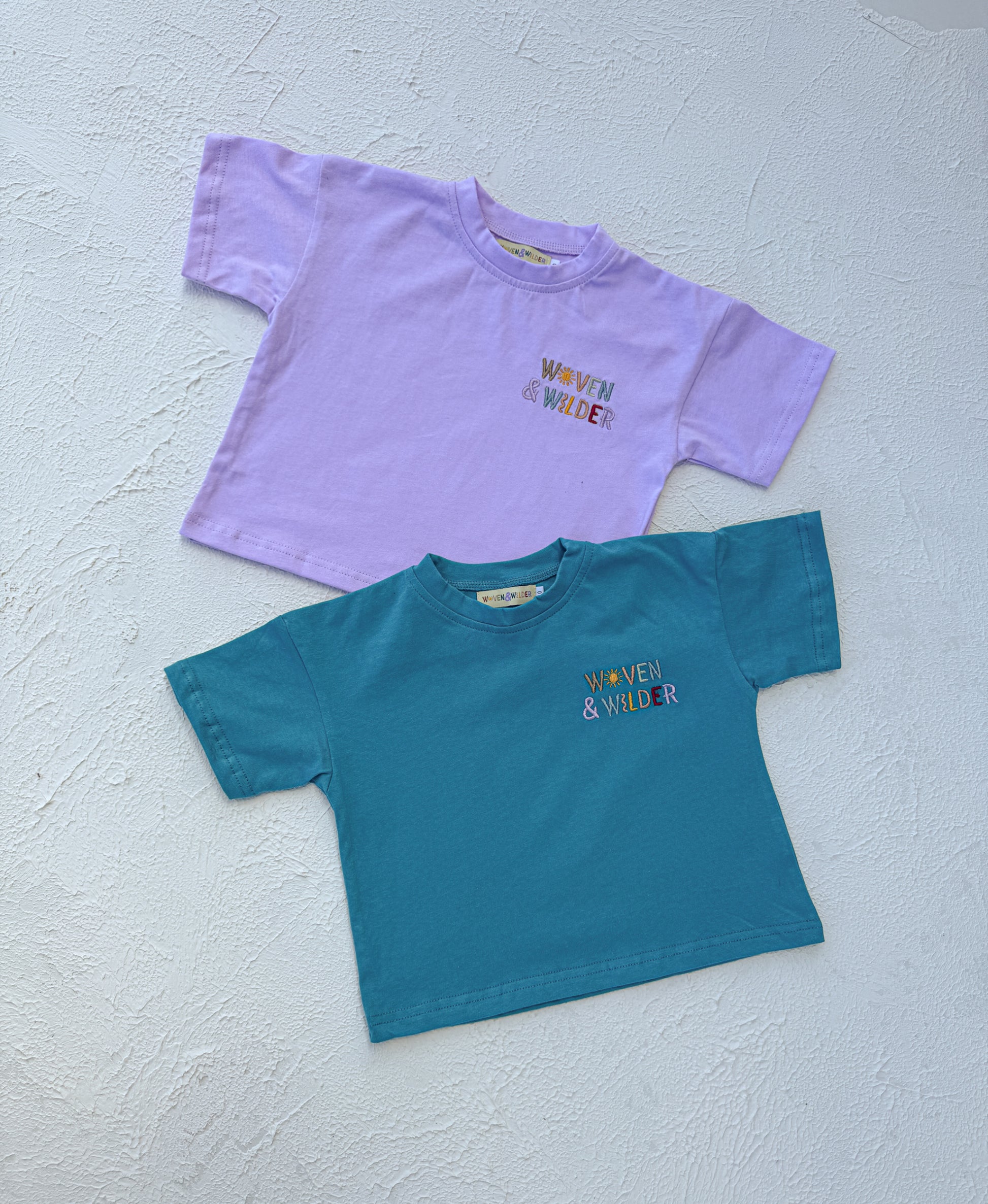 Two t-shirts, one purple and one blue, with 'Woven & Wider' text on a light gray background.