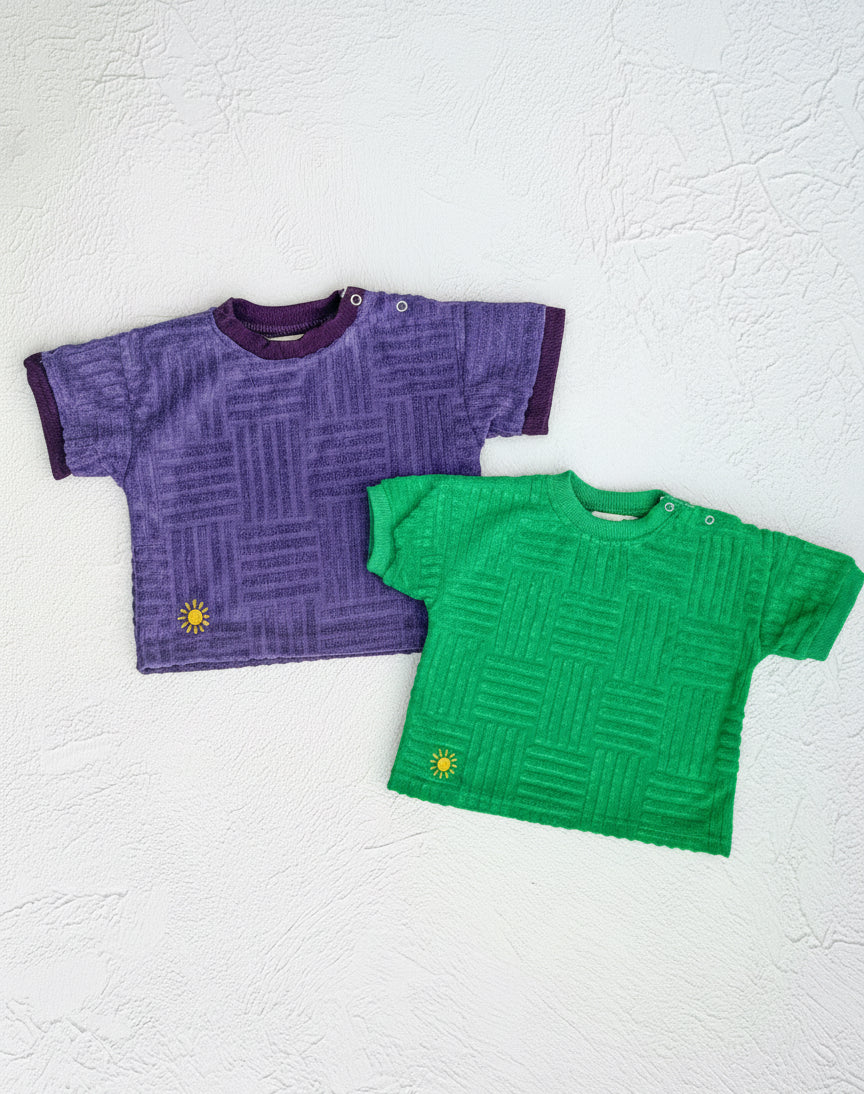 Two short-sleeve shirts, one purple and one green, on a textured white background.
