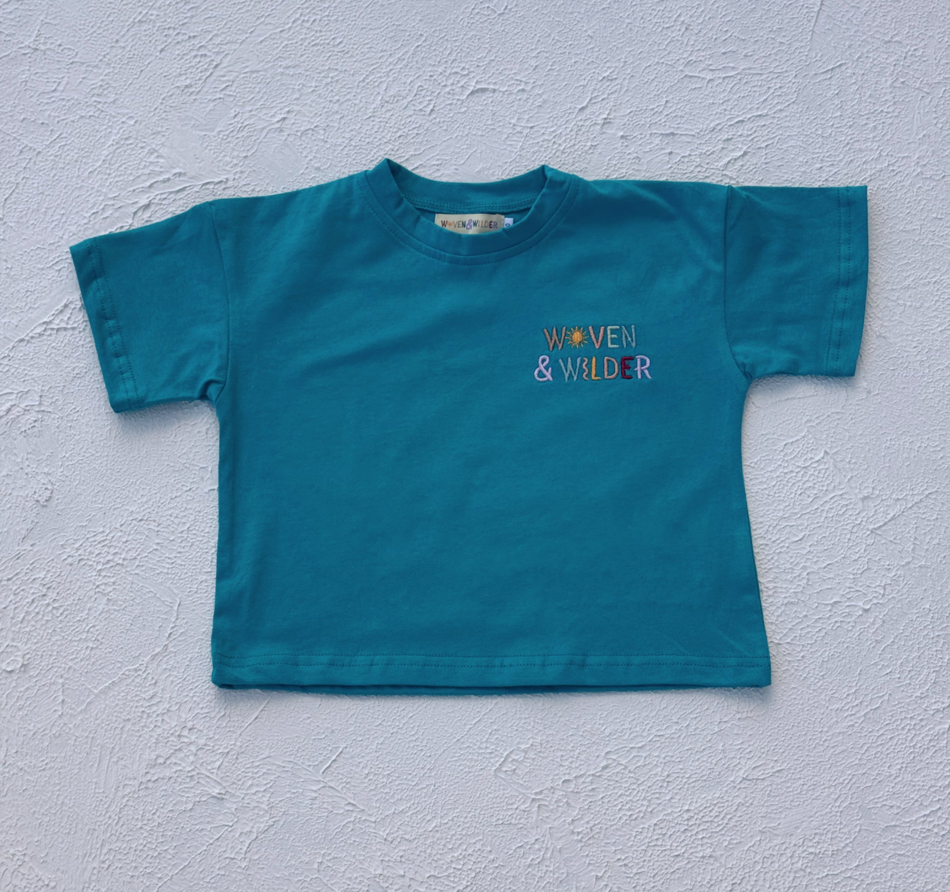 Blue t-shirt with 'Woven & Wilder' logo on a light gray background