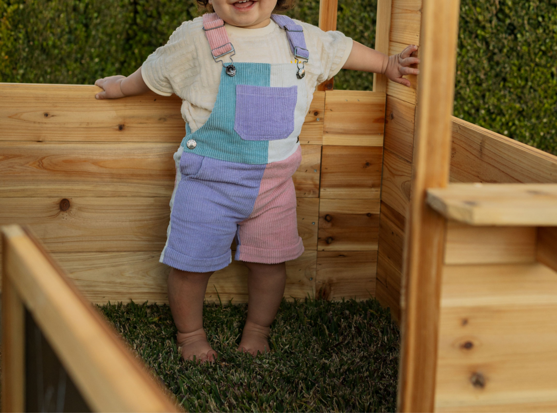 Child in a colorful overall standing inside a wooden playhouse.