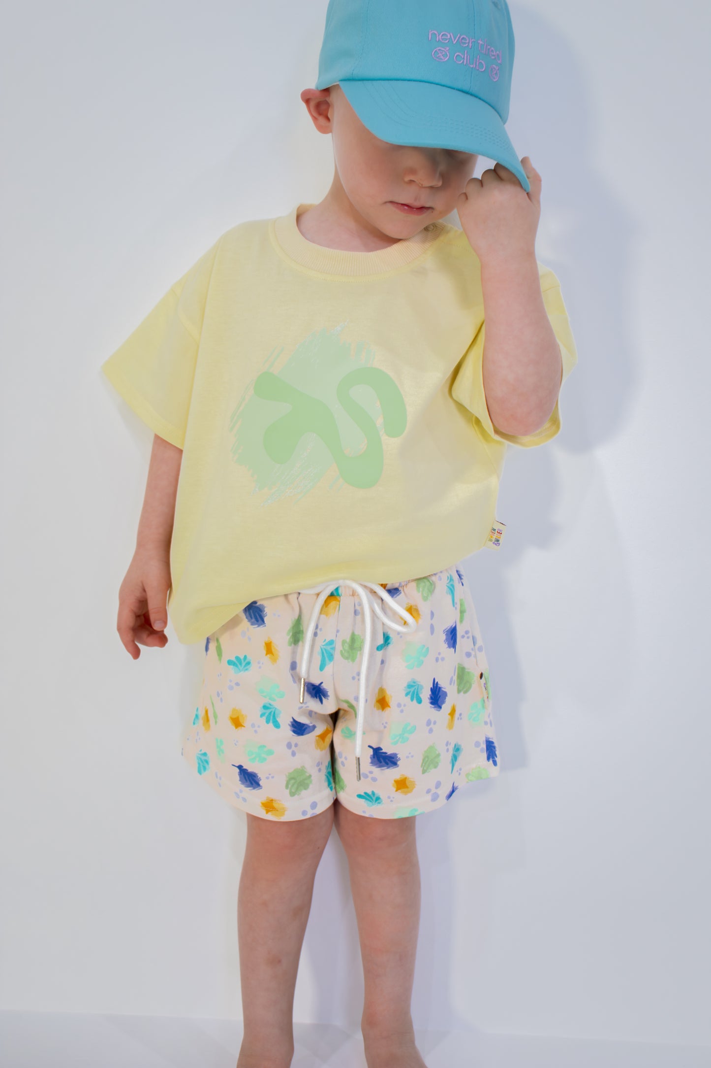 Child wearing a yellow shirt with a green graphic and patterned shorts on a white background