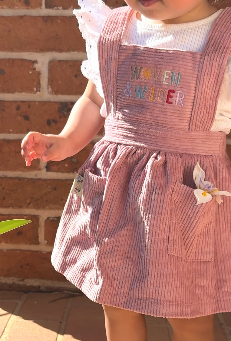 Child wearing a pink striped pinafore with embroidered text against a brick wall.