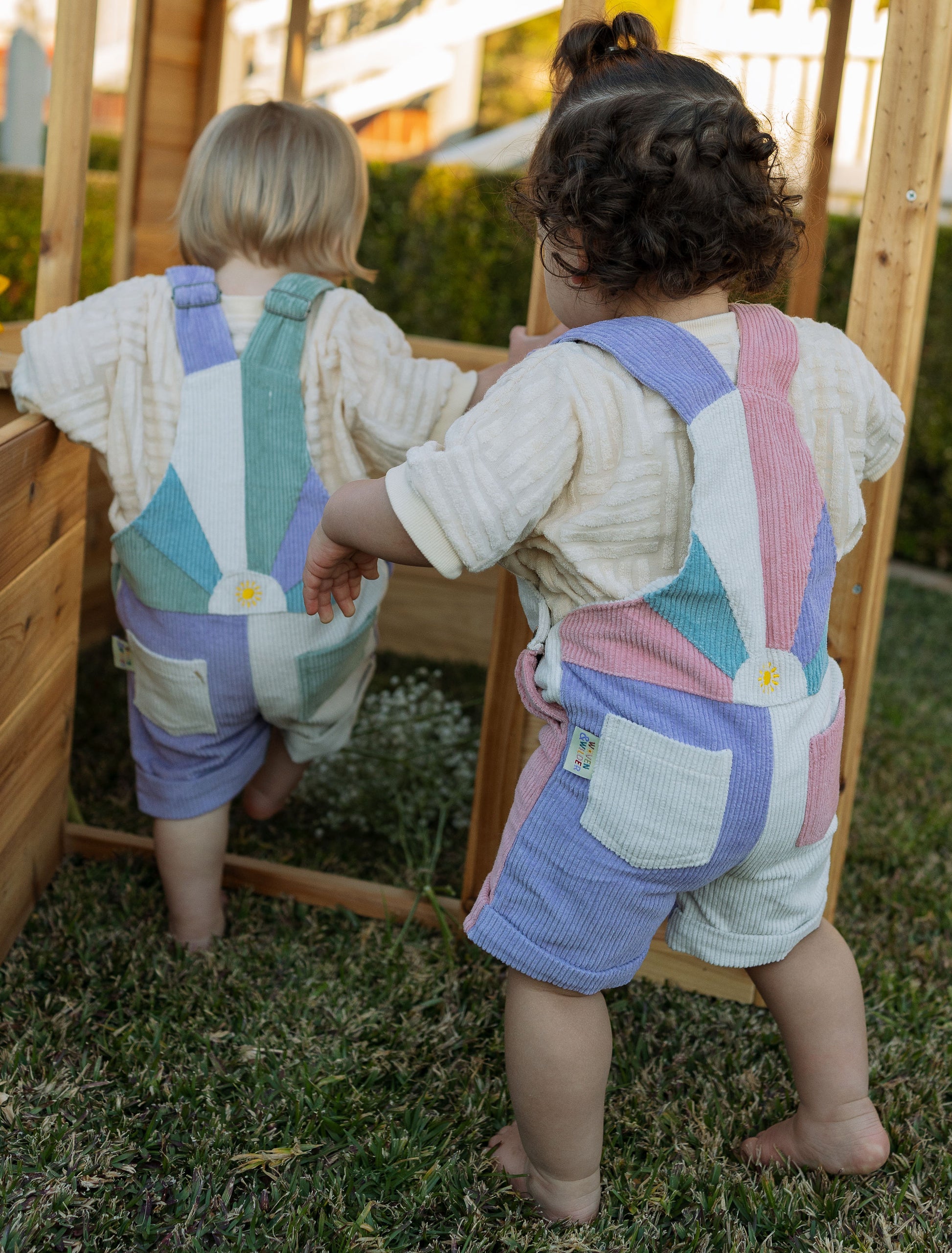 Two children wearing colorful overalls standing in a grassy area.