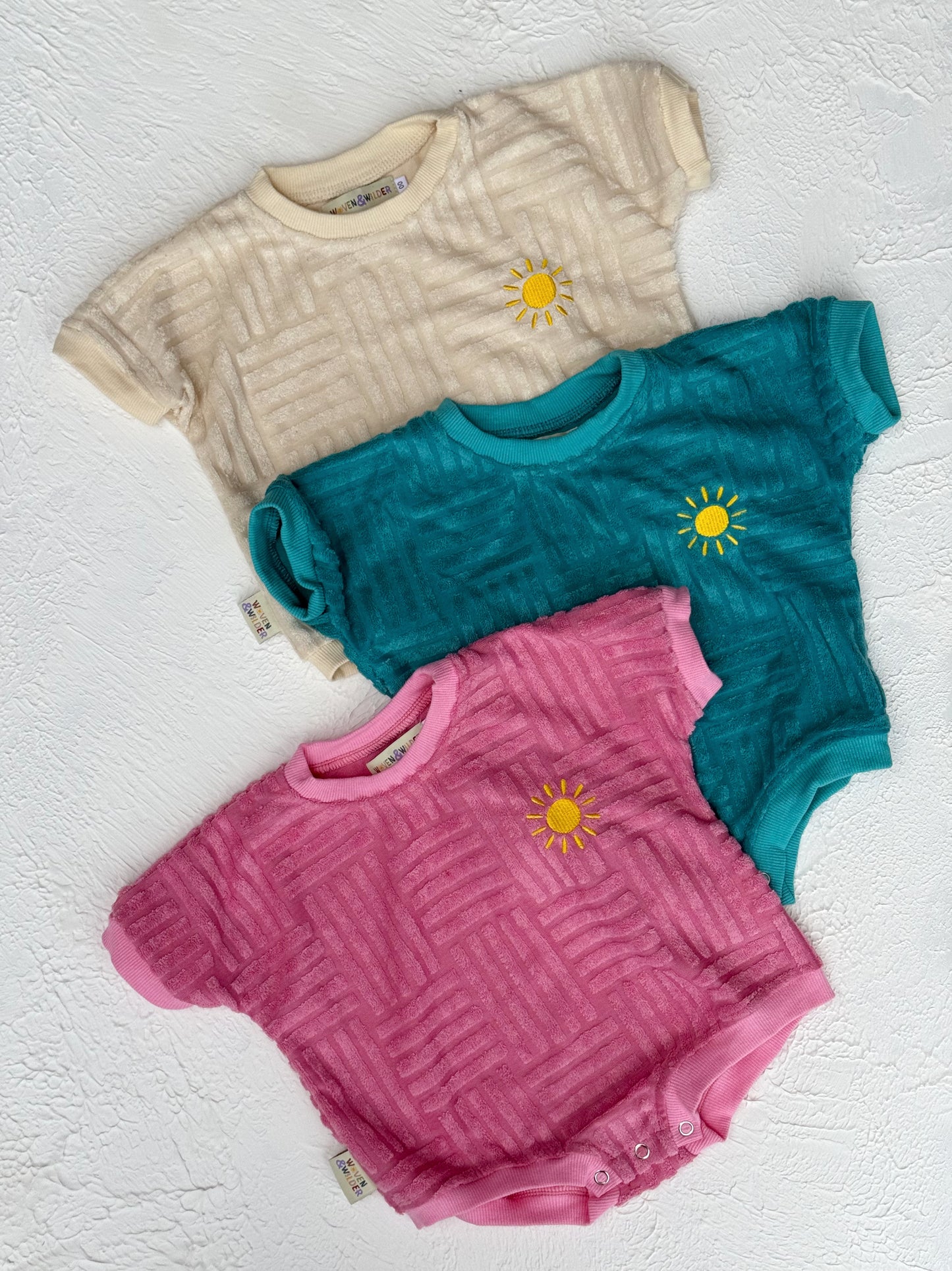 Three rompers in beige, teal, and pink with sun designs on a textured white surface.