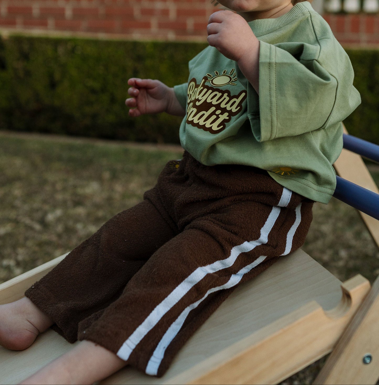 Child sitting on a wooden slide outdoors with a white picket fence in the background