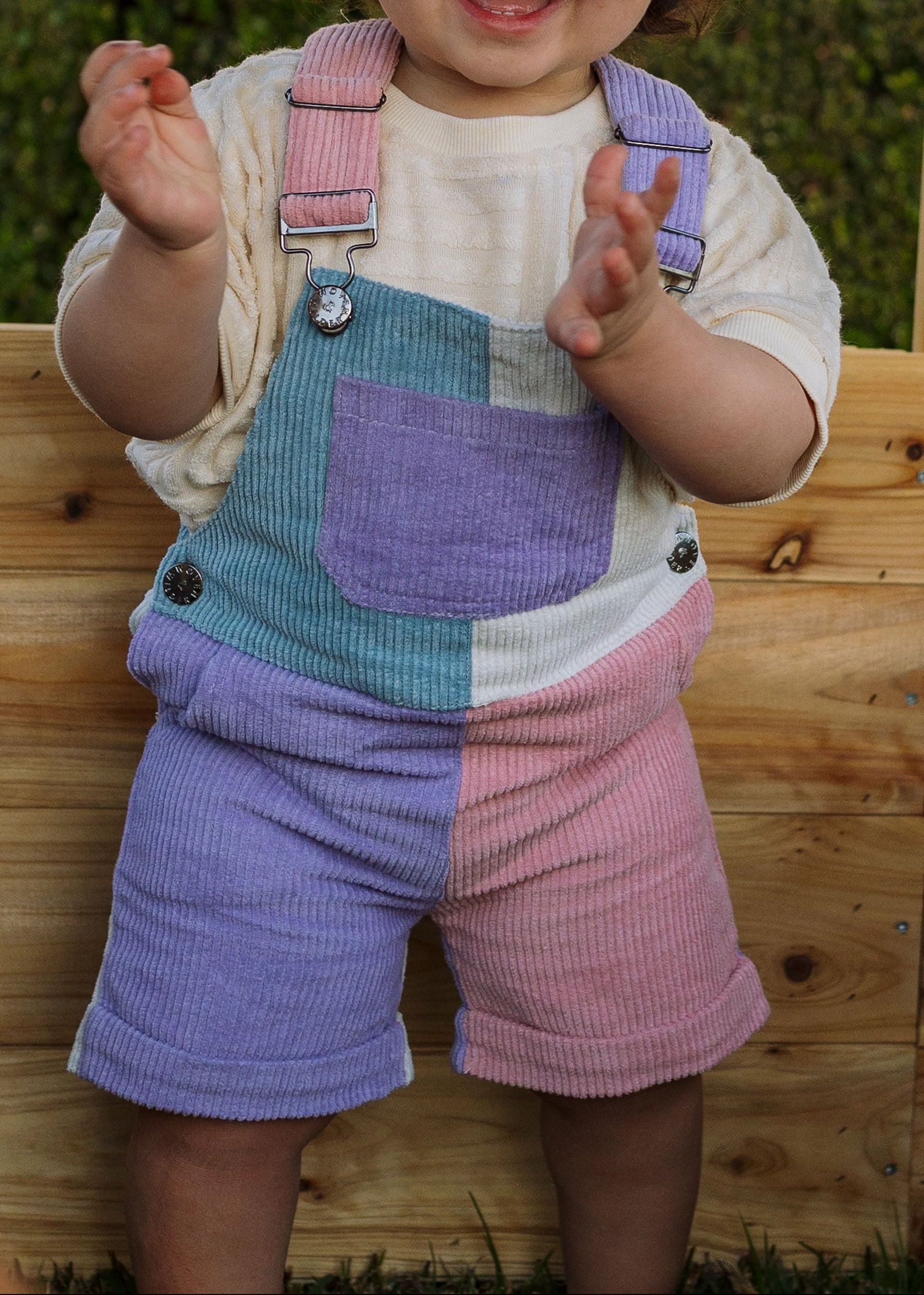 Child wearing colorful checkered overalls standing on a wooden bench with a green background