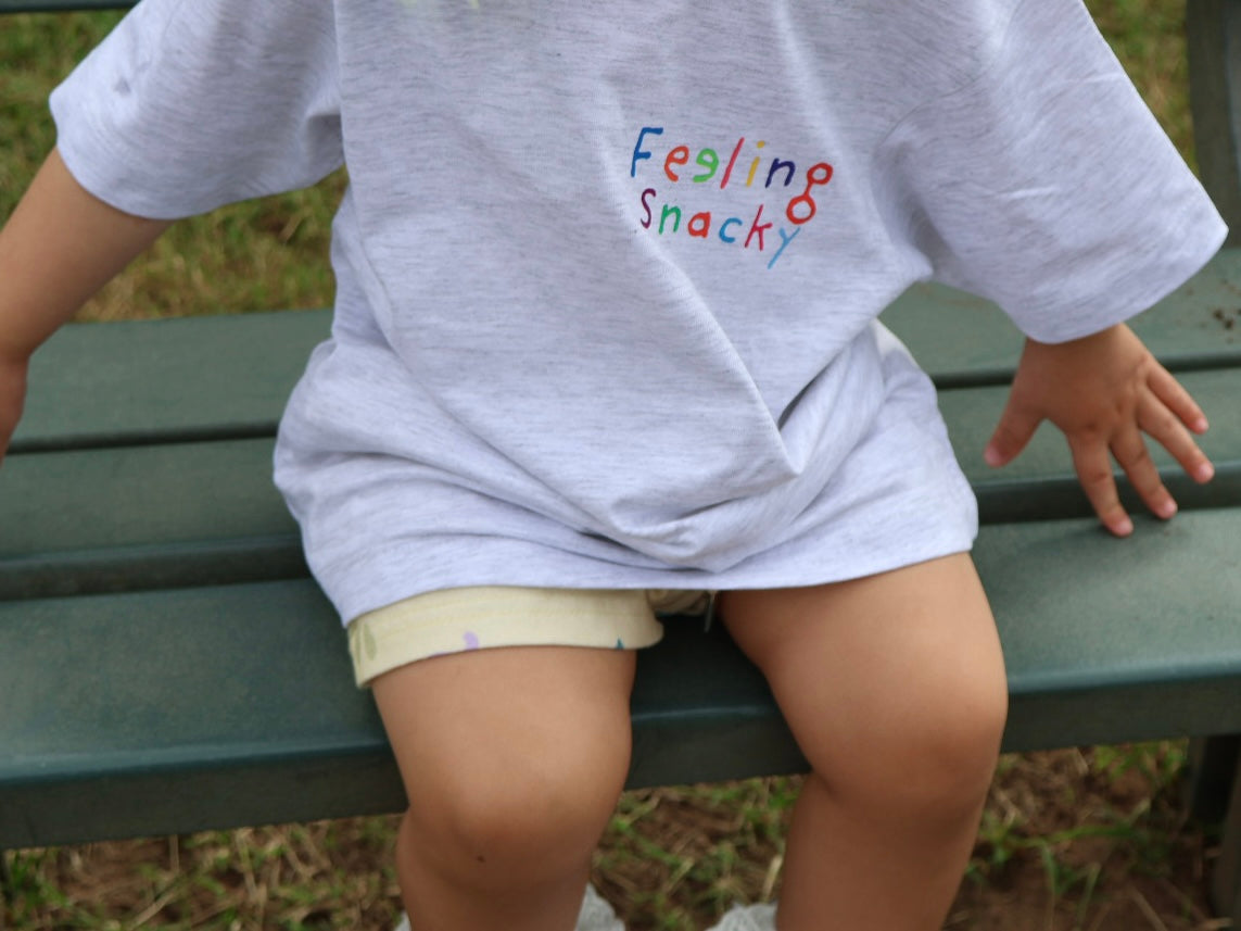 Child wearing a shirt with 'Feeling Snacky' text sitting on a bench.