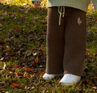 Child wearing a green sweater and brown pants standing in a grassy area with fallen leaves.